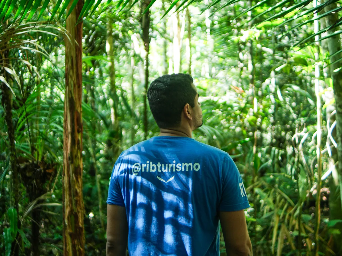 Viajante de camiseta azul da RIB olhando a mata fechada da Amazônia, sob folhas de palmeira
