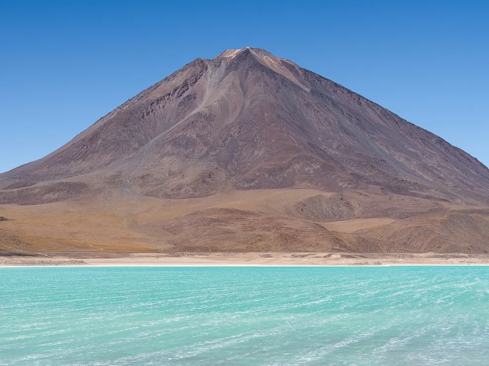 Vulcão de cume cônico diante de uma lagoa de água turquesa no altiplano boliviano, sob céu azul