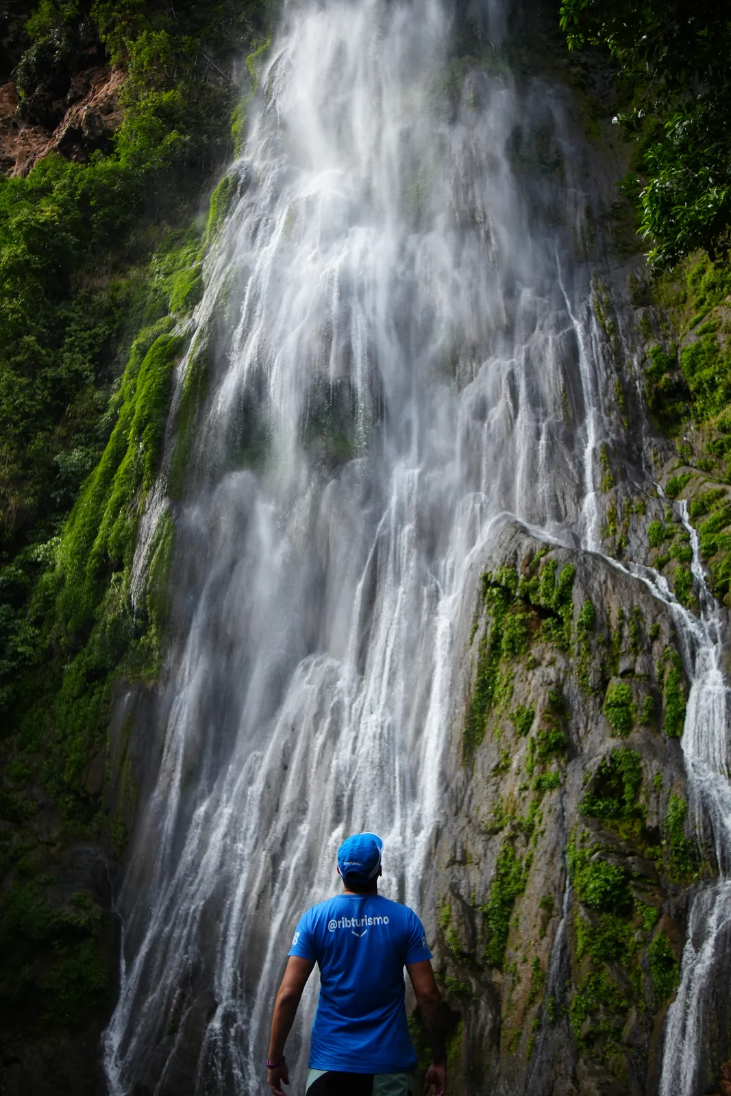 Guia da RIB de camiseta e boné azuis diante de uma cachoeira alta coberta de musgo