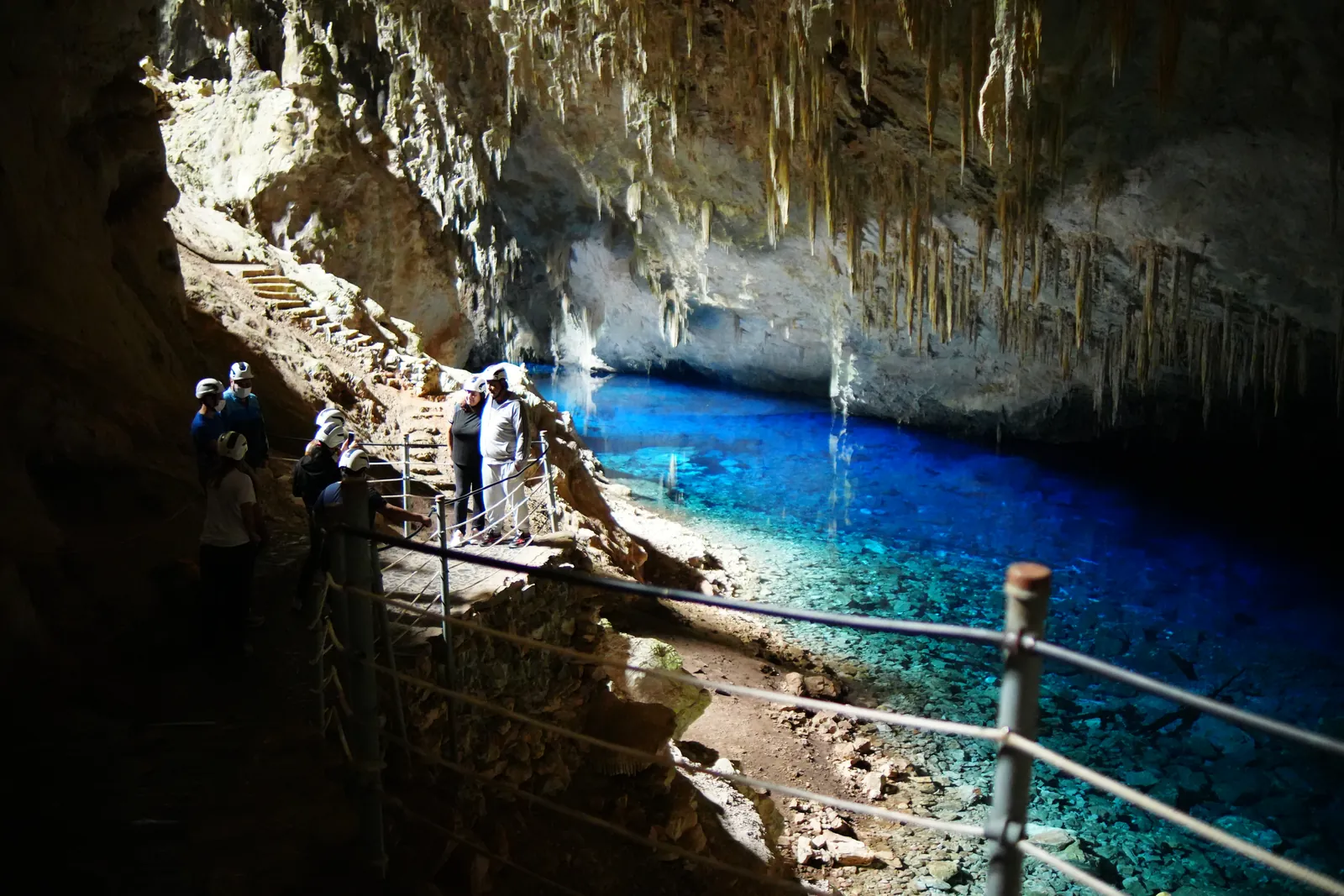 Viajantes de capacete numa passarela dentro de uma gruta, ao lado de um lago azul intenso