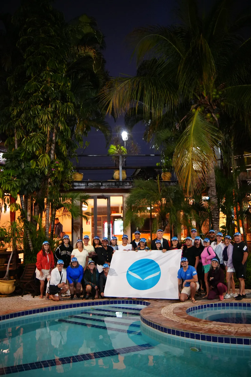 Grupo da expedição reunido à noite na beira da piscina, segurando a bandeira da RIB