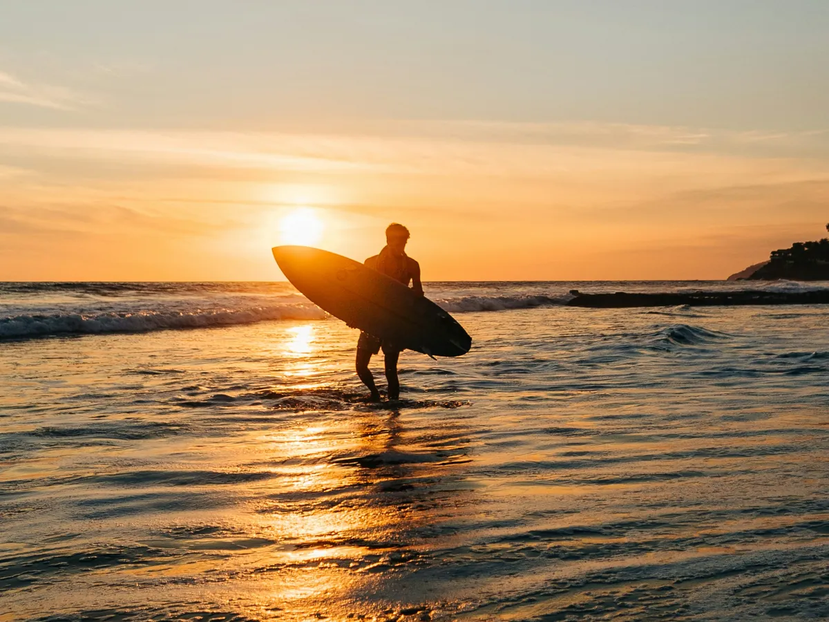Silhueta de surfista com a prancha saindo do mar em El Salvador, com o sol se pondo no horizonte