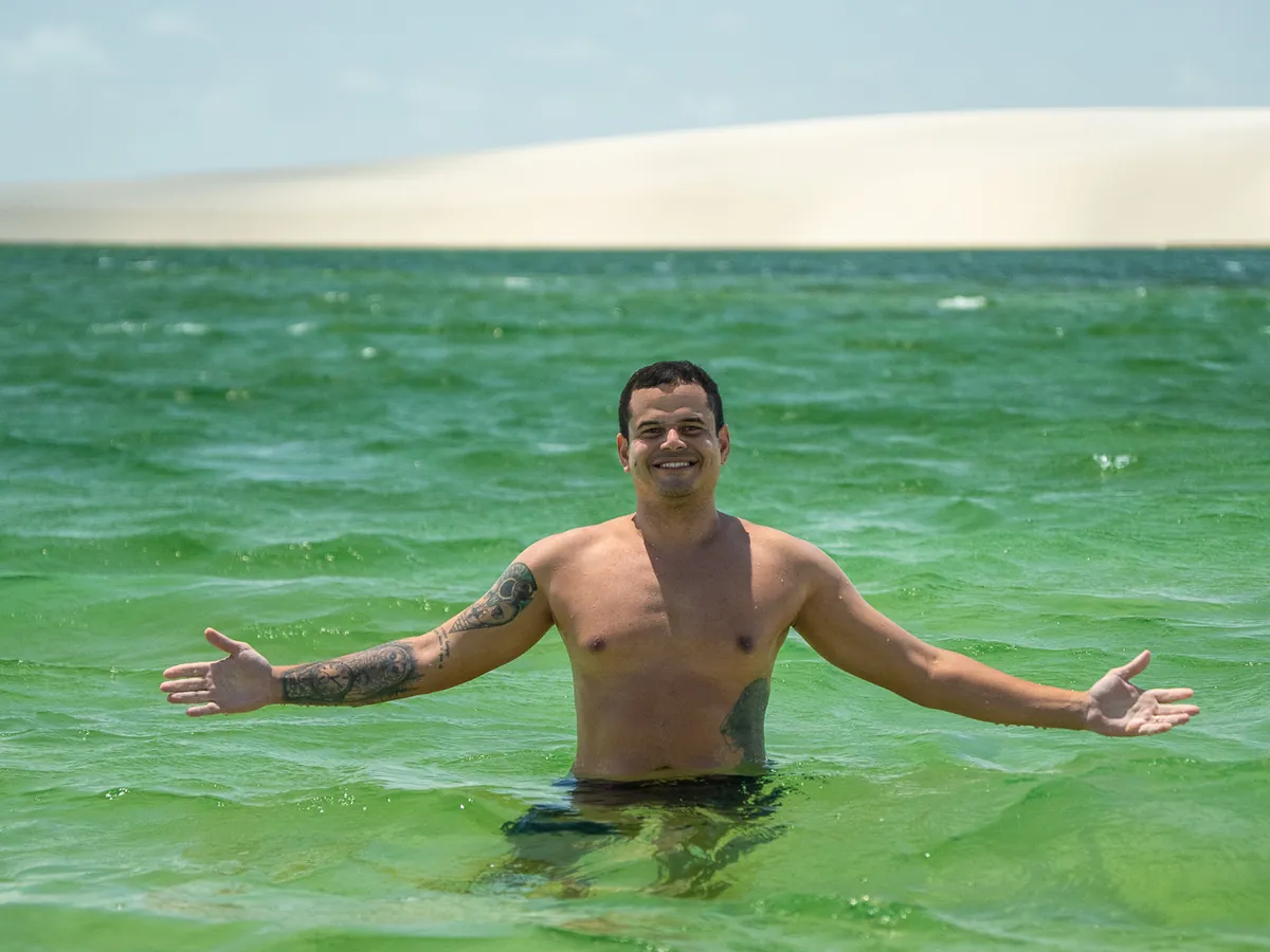 Viajante sorrindo de braços abertos numa lagoa verde, com uma duna branca ao fundo, nos Lençóis Maranhenses