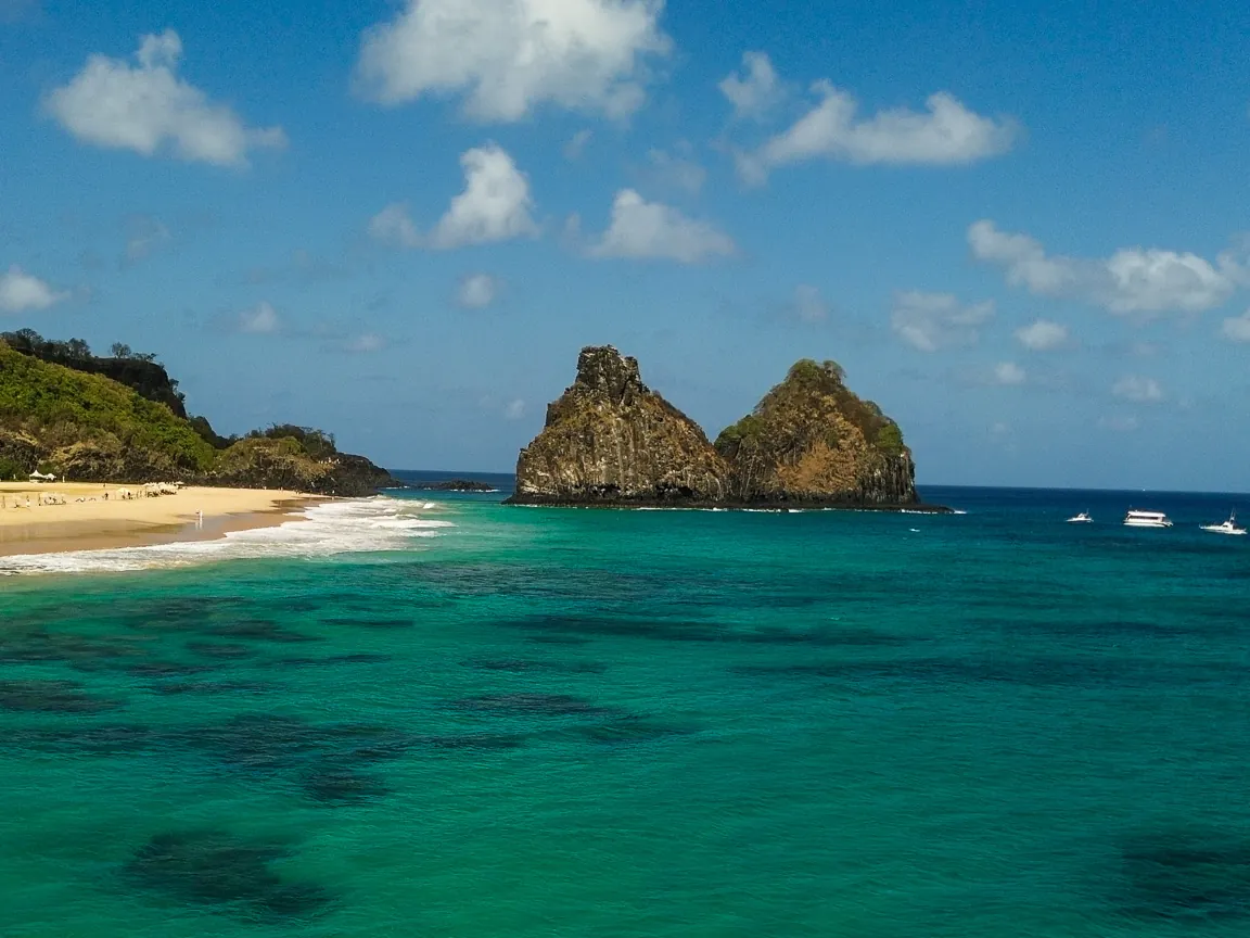 Morro Dois Irmãos saindo do mar verde-turquesa transparente, com praia de areia dourada e céu azul com nuvens em Fernando de Noronha